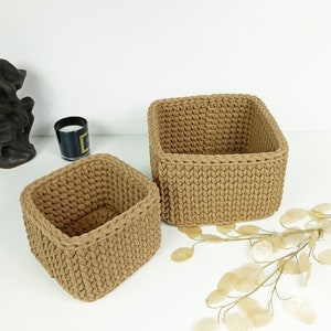 May include: Two square-shaped, brown, crocheted storage baskets. The baskets are made of a thick, textured yarn and have a woven pattern. The larger basket is in the background and the smaller basket is in the foreground.