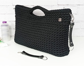 May include: A black crocheted handbag featuring a rectangular shape with integrated top handles, a detachable dark gray leather strap with silver-toned metal hardware, and a small rectangular brand tag labeled Agatka attached to the upper right side.