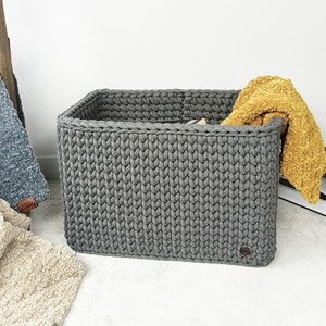 May include: A grey knitted storage basket with a yellow blanket inside. The basket is made of thick, chunky yarn and has a rectangular shape.