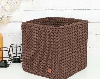 May include: A brown crocheted storage basket with a square shape. The basket is made of thick, textured yarn and has a subtle sheen.