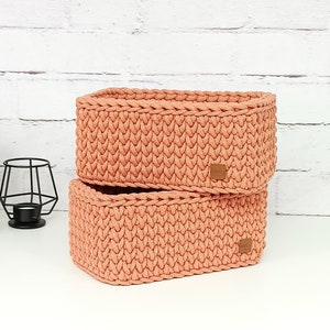 May include: Two rectangular storage baskets made of peach-colored yarn in a chunky knit pattern. Each basket features a small, square, brown leather-like tag attached to the front right side, displaying a subtle embossed logo design.
