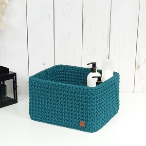 Puede incluir: Cesta de almacenamiento de crochet de color verde azulado con tres botellas blancas con bombas en su interior. La cesta está hecha de hilo grueso y tiene forma cuadrada.
