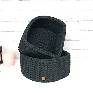 Puede incluir: Dos cestas de almacenamiento de crochet negras, una apilada sobre la otra. La cesta superior es ligeramente más grande que la cesta inferior. Ambas cestas tienen una forma cuadrada y una superficie texturizada.