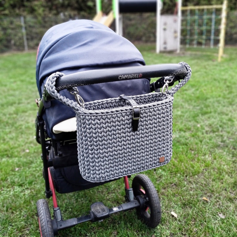 May include: A dark gray crocheted stroller organizer bag with a black leather strap and buckle closure. The bag features a braided gray cord handle attached to the black handlebar of a navy blue stroller with the brand name CAMARELO printed on it.