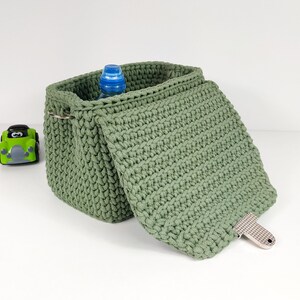 May include: A green crocheted storage basket with a lid. The basket is open and a blue plastic bottle is inside.