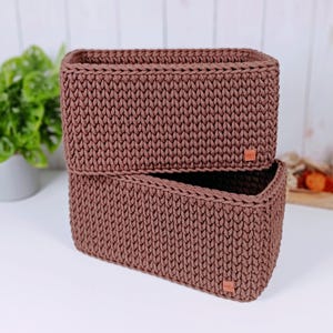 Peut inclure: Deux paniers de rangement rectangulaires en corde crochetée marron. Chaque panier présente un motif en maille uniforme et une petite étiquette carrée en similicuir marron clair fixée sur le coin inférieur droit de la face avant.