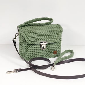 May include: A green crocheted handbag with a brown leather strap and a silver clasp. The bag has a flap closure and a small pocket on the front.