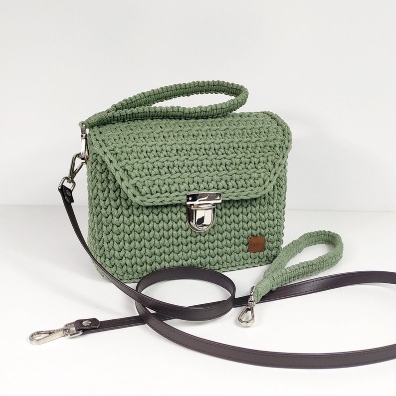 May include: A green crocheted handbag with a brown leather strap and a silver clasp. The bag has a flap closure and a small pocket on the front.