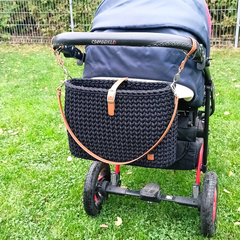 May include: A black stroller with a black crocheted bag attached to the handle. The bag has a brown leather strap and buckle. The stroller has a black fabric seat and a black handlebar with the word "CAMARELO" printed on it.
