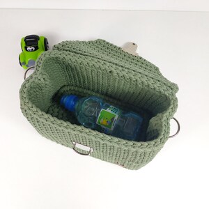 May include: A green crocheted storage basket with a metal buckle and two metal rings. The basket is open and a blue plastic water bottle is inside.