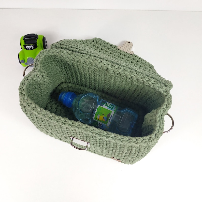 May include: A green crocheted storage basket with a metal buckle and two metal rings. The basket is open and a blue plastic water bottle is inside.
