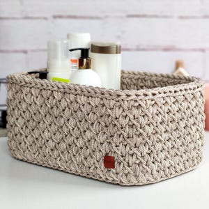 Op de afbeelding: Een beige, rechthoekige, gehaakte mand met een textuurpatroon. De mand is gevuld met witte flessen en containers. Een klein, bruin label is aan de voorkant van de mand bevestigd.