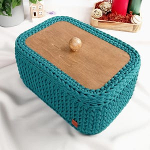 Op de afbeelding: Een rechthoekige turquoise gehaakte opbergmand met een houten deksel. Het deksel heeft een kleine houten knop. De mand is gemaakt van dik, getextureerd garen. Het algehele ontwerp is handgemaakt en decoratief.