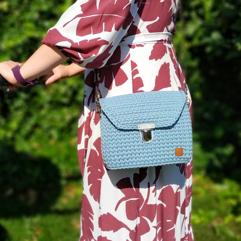 May include: A light blue crocheted crossbody bag with a silver clasp and a brown leather patch. The bag is worn by a person wearing a white and burgundy floral dress.