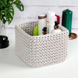 May include: A square, light gray, crocheted basket filled with various white and beige toiletries. The basket is sitting on a white surface, with a green plant and a green candle in the background. The basket is made of thick yarn.