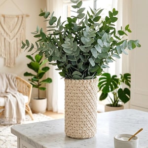 Può includere: Un vaso alto e cilindrico in macramè beige contiene una grande composizione di rami di eucalipto fresco con foglie verdi e verde-grigio. Il vaso è appoggiato su un piano di lavoro in marmo bianco, creando un'estetica naturale e neutra.