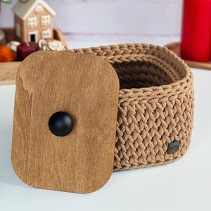 Peut inclure: Un panier de rangement rond en corde crochetée de couleur beige avec un couvercle en bois brun clair lisse. Le couvercle est doté d'un bouton noir rond centré. Une petite étiquette rectangulaire noire est fixée sur le côté du panier.