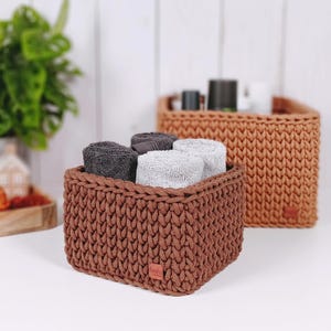 May include: Two brown, rectangular, woven baskets. The front basket contains rolled gray and white towels. The second basket is in the background and contains toiletries. The baskets are on a white surface.