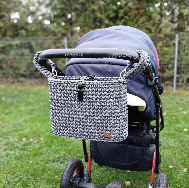 May include: A dark gray crocheted stroller organizer bag with a black leather-like strap and buckle closure. The rectangular bag features a textured knit pattern and is attached to the black handle of a navy blue stroller with two gray braided cords.
