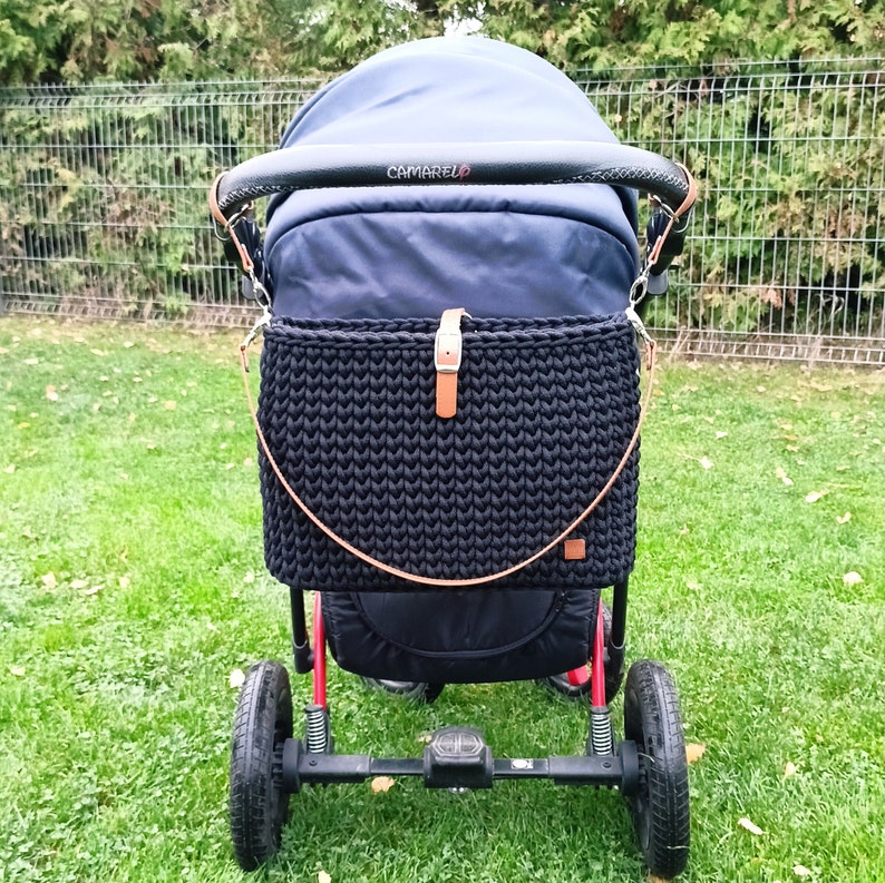 May include: A black stroller with a black crocheted bag attached to the handle. The stroller has a black canopy with the word "CAMARELO" printed on it.