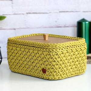May include: A handmade, yellow-green crochet basket with a wooden lid. The basket is rectangular with rounded corners and a small wooden knob on top. A small brown tag is attached to the front of the basket.