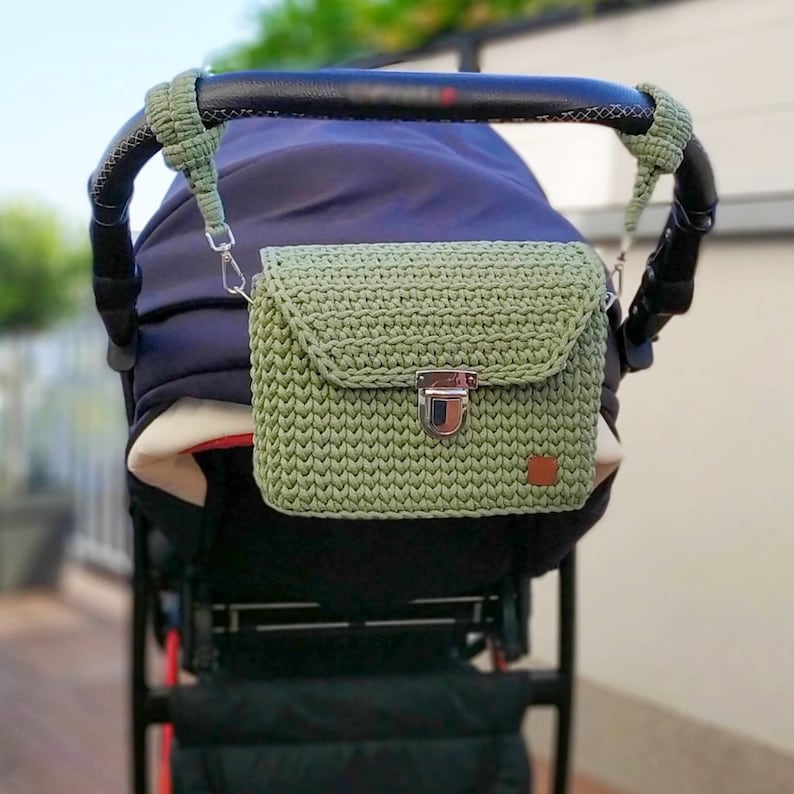 May include: A green crocheted bag with a silver clasp attached to a stroller handle.