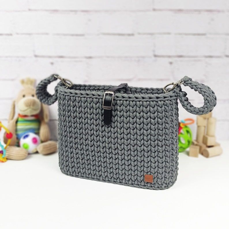 May include: A rectangular dark gray crocheted bag featuring a thick, textured knit pattern. The bag includes a black faux leather strap with a silver metal buckle closure at the top center and a small brown rectangular brand tag at the bottom right.