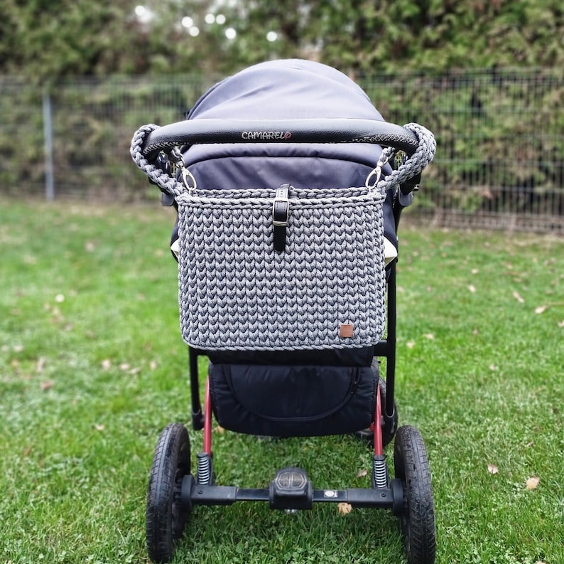 May include: A dark gray crocheted rectangular bag with a black leather strap and buckle closure, attached to the handlebar of a black stroller. The bag features a textured knit pattern and a small brown rectangular tag on the bottom right corner.
