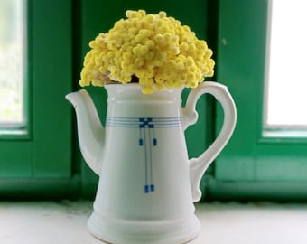 May include: A white ceramic teapot-shaped vase with blue geometric designs, filled with bright yellow, spherical flowers. The vase sits on a white surface in front of a green window frame.