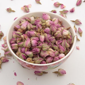 Op de afbeelding: Een witte kom gevuld met gedroogde roze rozenknoppen. De kom heeft een bloemenpatroon en is omringd door losse rozenknoppen op een wit oppervlak.