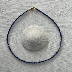 May include: A delicate necklace made of small, dark blue beads. The necklace is on a white surface.