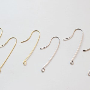 May include: Six earring hooks, three gold and three silver, are arranged in a row on a white surface. The hooks are all the same size and shape, with a small loop at the top and a long, thin wire that curves into a hook.