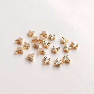 Puede incluir: Una colección de cuentas doradas en forma de mariposa. Las cuentas están esparcidas sobre una superficie blanca.