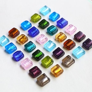 Puede incluir: Una colección de cuentas de vidrio cuadradas y coloridas en varios tonos de azul, verde, rosa, amarillo y marrón. Las cuentas están dispuestas en un patrón de cuadrícula, mostrando su cualidad translúcida y superficies reflectantes. Ideal para la fabricación de joyas o proyectos de manualidades.