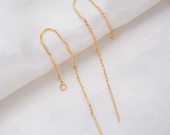 May include: A pair of gold-colored threader earrings. Each earring features a curved top, a delicate chain, and a straight bar at the end. The earrings are displayed on a white, textured surface, highlighting their elegant design.