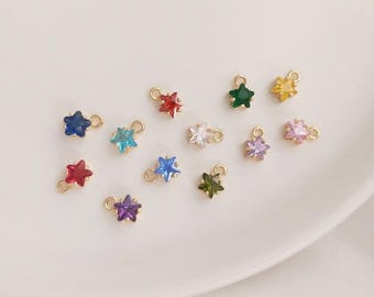 May include: A collection of small star-shaped charms in various colors, including blue, red, green, and yellow. Each charm has a gold-colored loop for attachment. The charms are displayed on a white surface.