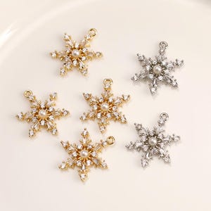Op de afbeelding: Vijf sneeuwvlokcharms met een parel in het midden. Twee charms zijn goud en drie zijn zilver.