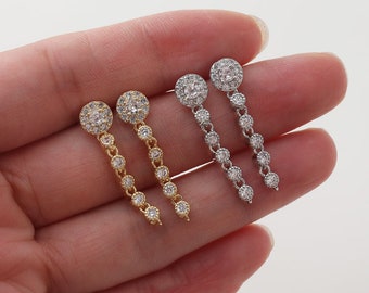 May include: A pair of dangle earrings with a round, clear crystal center surrounded by smaller crystals. The earrings are available in gold and silver.