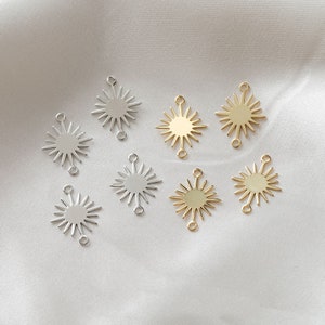 Op de afbeelding: Acht gouden en zilveren zonnestralencharms met elk twee springringen. De charms zijn gerangschikt in twee rijen op een witte stoffen achtergrond.