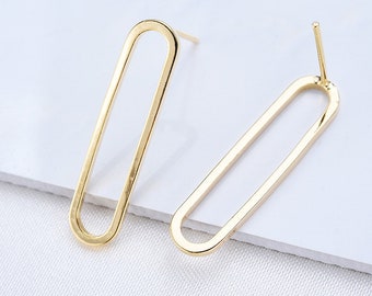 Puede incluir: Un par de pendientes dorados con marco abierto. Cada pendiente presenta una forma rectangular larga y delgada con extremos redondeados. Los pendientes se muestran sobre una superficie blanca, mostrando su diseño minimalista y brillo metálico.