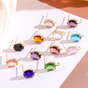 May include: A selection of stud earrings featuring gold-tone settings and oval-cut gemstones in a variety of colours. The gemstones include clear, pink, purple, blue, green, orange, and black. Each earring has a small loop detail.