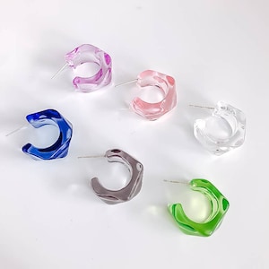 May include: Five pairs of colorful, geometric hoop earrings. The earrings are made of clear plastic and have a slightly irregular shape. The colors are pink, blue, clear, gray, and green.