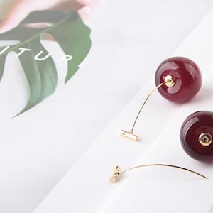 Puede incluir: Un par de pendientes de oro con abalorios en forma de cereza roja. Las cerezas son de vidrio y tienen un acabado brillante.