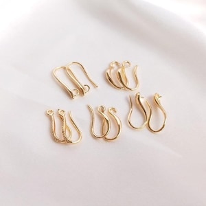 Può includere: Un set di sei ganci per orecchini dorati, alcuni con un design decorativo. I ganci sono disposti in tre coppie su una superficie bianca.