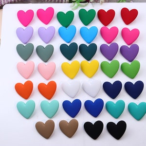 May include: A set of 24 heart-shaped buttons in various colors, including pink, purple, blue, green, yellow, orange, red, white, black, brown, and turquoise. The buttons are arranged in rows on a white surface.