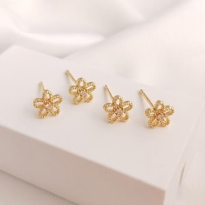 May include: Four gold-colored flower stud earrings with a small, clear gem in the center of each flower. The earrings are displayed on a white surface, with a soft, white background.