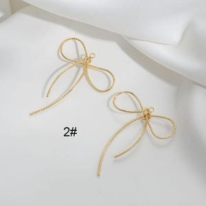 Peut inclure: Deux breloques d'oreille en forme de nœud en métal doré avec une surface texturée. Les breloques sont présentées sur un fond blanc. 2#