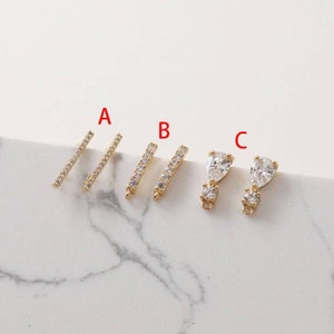 Puede incluir: Componentes de joyería dorados, que incluyen dos pares de barras lineales con cristales transparentes incrustados y un par de colgantes en forma de lágrima con un cristal más pequeño debajo. Los componentes están etiquetados A, B y C.