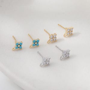 May include: A collection of stud earrings in gold and silver tones. Some earrings feature a cluster of small, turquoise-colored stones, while others have clear, sparkling crystals. The earrings are arranged on a light-colored surface.