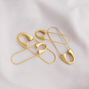 Può includere: Un paio di orecchini a forma di spilla di sicurezza dorati. Gli orecchini sono in metallo e hanno un design semplice e minimalista.